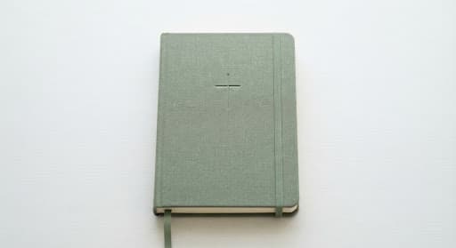 Carnet relié en lin vert sauge avec croix minimaliste embossée, photographié de dessus sur fond blanc